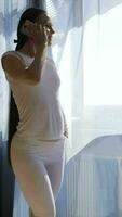 a pregnant woman in white pants and a white shirt standing by the window is using her phone video