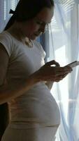 a pregnant woman in white pants and a white shirt standing by the window is using her phone video