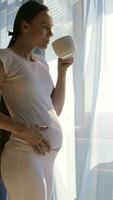 a pregnant woman in white pants drinking coffee while standing by the window video