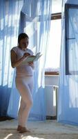 a pregnant woman in white pants and a white shirt is standing in front of a window reading a book video