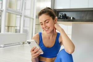 Image of young fit woman wearing sportbra and blue leggings, using smartphone app, does workout from home, sits on floor and follows online training video photo