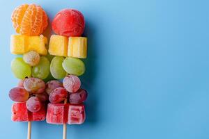 A vibrant assortment of frozen fruits on skewers, including oranges, watermelon, pineapple, grapes, and strawberries, against a blue background photo