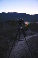 Vertical shot of modern digital camera placed on tripod in nature outdoors, ready for capturing steady pictures or of a village in mountains at dawn. World photography day concept. photo