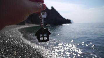 Keys with a house-shaped keychain in a man's hand on sea beach background. The concept of buying a home, apartment, real estate, young family, home loan, rental housing. Close-up, Slow motion video