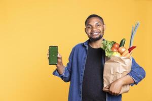 African american guy shows greenscreen display on smartphone, carrying a paper bag with bio produce against yellow background. Male model uses mobile phone with isolated mockup. photo