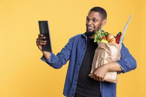 Smiling male model uses his tablet to check socials in studio, holding a bag full of vegetables and fruits. Confident young adult looks at device and laughing against yellow background. photo