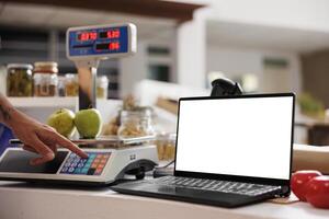 Digital laptop displaying white screen is positioned on checkout counter of eco friendly store. While storekeeper weighs apples on scale, the wireless computer shows blank copyspace mockup template. photo