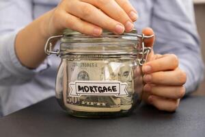 Unrecognizable woman holding Saving Money In Glass Jar filled with Dollars banknotes. MORTGAGE transcription in front of jar. Managing personal finances extra income for future insecurity background photo