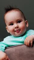 Little adorable baby stands on the chair looking down at camera. Nice Caucasian kid showing tongue and waving hand. Low angle view. Vertical video