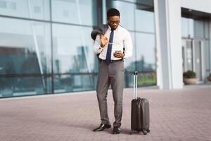 African American businessman is making a call by the entrance of a modern airport, with luggage beside him. The mood is professional and active, showcasing travel and communication. photo