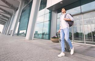 Homeland Arrival Concept. Joyful millennial guy walking out of airport entrance with luggage, low angle view, free space photo