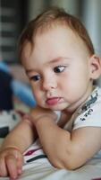 Little sweet calm baby boy near the bed. Adorable Caucasian child looking into camera with interest. Close up. Blurred backdrop. Vertical video