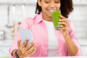 Closeup portrait of black girl looking at cell phone, drinking green healthy vegetable juice, selective focus photo