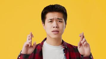 Asian young man stands with his eyes closed and fingers crossed, symbolizing hope and luck. The bright yellow background adds a cheerful and optimistic vibe to the moment. video