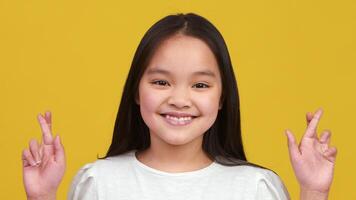 A cheerful Asian girl with long hair smiles broadly while crossing her fingers, expressing hope or good luck against a bright yellow backdrop. video