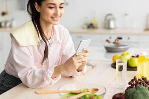 Modern Technologies And Lifestyles Concept. Portrait of cheerful young female using cellphone while leaning on kitchen countertop, smiling maid reading text messages on her cellphone, browsing web photo