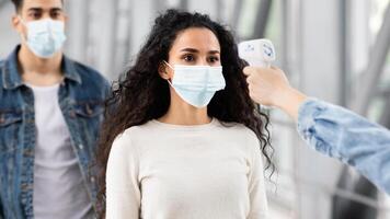 Safe Travels. Staff Checking Passengers Temperature With Electronic Thermometer At Airport Entrance For Covid-19 Outbreak Prevention, Middle Eastern Couple In Medical Masks Waiting Turn To Enter photo