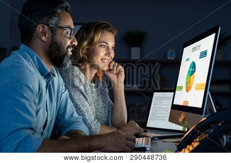 Happy business people working together in office at night. Startup business partners working late together. Mature man and casual woman working with computer late at night in their office. 