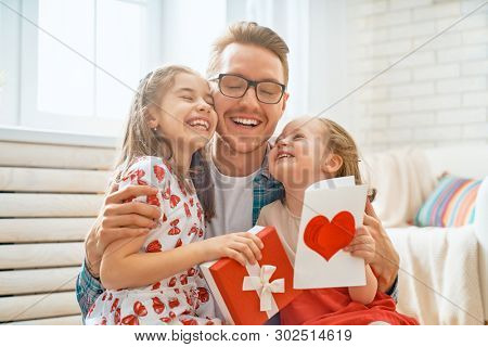 Happy father's day! Children daughters congratulating dad and giving him gift box. Daddy and girls smiling and hugging. Family holiday and togetherness.