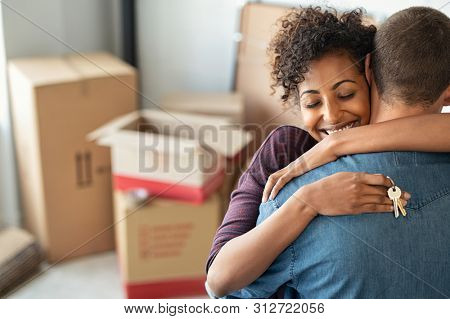 Young african woman holding home keys while hugging boyfriend in their new apartment after buying real estate. Lovely girl holding keys from new home and embracing man. Couple around cardboard boxes.