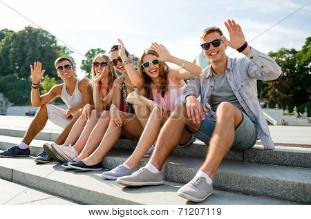 friendship, leisure, summer, gesture and people concept - group of smiling friends sitting on city street and waving hands