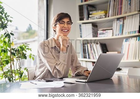 Successful young business woman sitting in creative office and looking at camera. Portrait of happy entrepreneur working on computer. Smiling businesswoman using laptop while working from home.