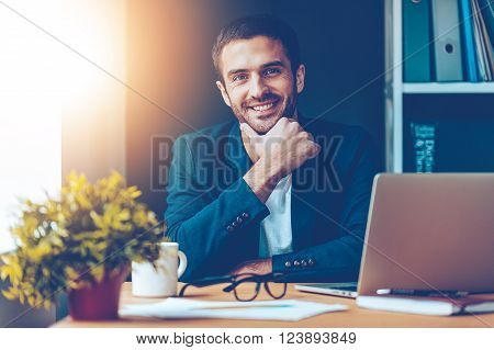 Confident and handsome. Confident young man holding hand on chin and smiling while sitting at his working place in office