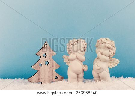 Two Christmas Cherub Baby Angels Statuettes On Snow With Wooden Christmas Tree On Light Blue Backgro