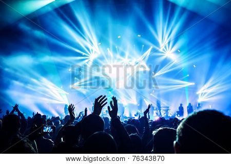 Large group of happy people enjoying rock concert, clapping with raised up hands, blue lights from the stage, new year celebration concept