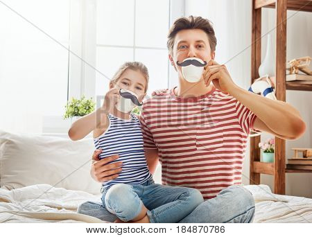 Happy father's day! Dad and his child daughter are playing and having fun together. Beautiful funny girl and daddy have mustaches on cups. Family holidays and togetherness.