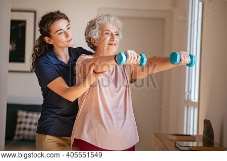 Old woman training with physiotherapist using dumbbells at home. Therapist assisting senior woman with exercises in nursing home. Elderly patient using dumbbells with outstretched arms.