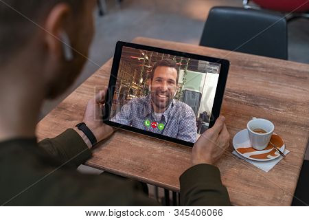 Smiling business partners talking through video call. Back view of man in coffe shop doing video chat calling a friend. Young man and his friend talking to each other through a videocall on tablet.