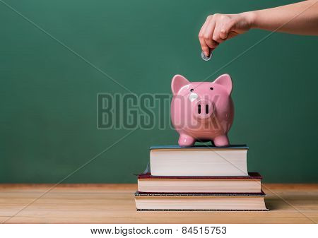 Person Depositing Money In A Piggy Bank On Top Of Books With Chalkboard