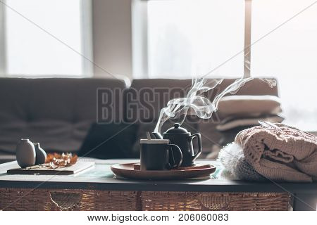 Still life details in home interior of living room. Sweaters and cup of tea with steam on a serving tray on a coffee table. Breakfast over sofa in morning sunlight. Cozy autumn or winter concept.