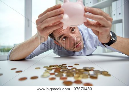 business, people, finances, crisis and money saving concept - businessman shaking piggy bank and getting coins at office