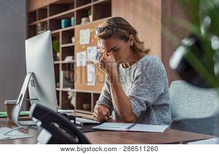 Exhausted businesswoman having a headache at office. Mature creative woman working at office desk feeling tired. Stressed casual business woman feeling eye pain while overworking on desktop computer.