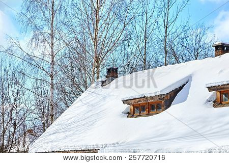 Roof Of A Low-rise Wooden Village House With Windows Under The Snow. Winter. Trees And Blue Sky With