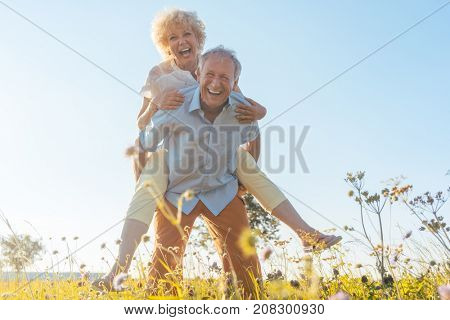 Low-angle view portrait of a happy senior man laughing while carrying his partner on his back, in a sunny day of summer in the countryside