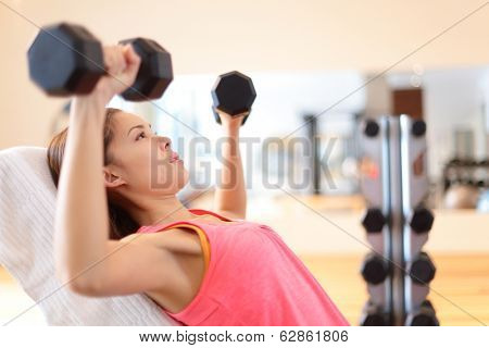 Gym woman strength training lifting dumbbell weights in shoulder press exercise. Female fitness girl exercising indoor in fitness center. Beautiful fit mixed race Asian Caucasian model training.