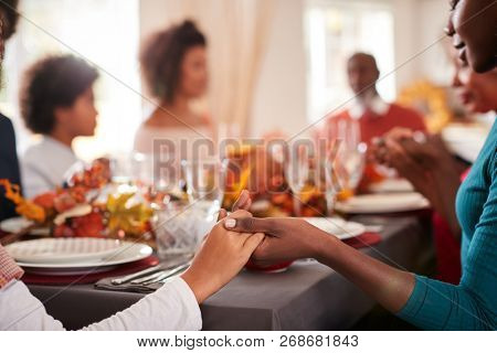 Young black adult woman and her daughter holding hands and saying grace with their multi generation family at the Thanksgiving dinner table, detail, focus on foreground