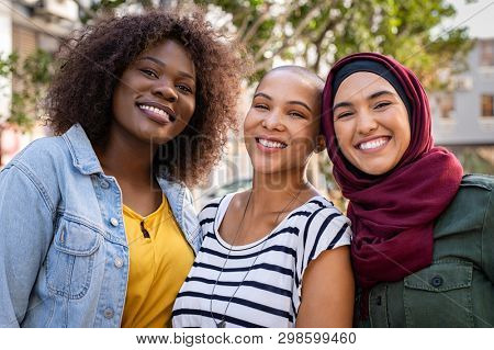 Group of three happy multiethnic friends looking at camera. Portrait of young women of different cultures enjoying vacation together. Smiling islamic girl with two african american friends outdoor.