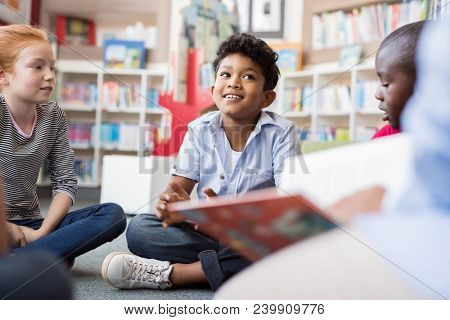 Multiethnic group of kids sitting on floor in circle around the teacher and listening a story. Discussion group of children inaa library talking to woman. Smiling hispanic boy in elementary school.