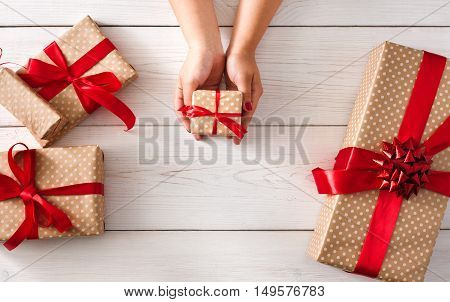 DIY hobby. Woman's hands give wrapped christmas holiday handmade present in paper with red ribbon. Present box, decoration of gift on white wooden table, top view with copy space