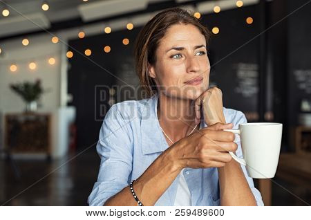 Thoughtful mature woman sitting in cafeteria holding coffee mug while looking away. Middle aged woman drinking tea while thinking. Relaxing and thinking while drinking coffee.