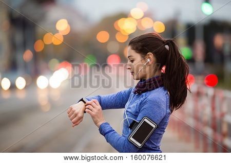 Beautiful young woman in the city with smart phone, smartwatch and earphones, listening music. Using a fitness app for tracking weight loss progress, running goal or summary of her run. Rear view.