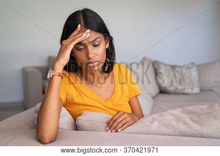 Sad depressed indian woman at home sitting on couch. Stressed young woman alone at home with copy space. Frustrated and unhappy middle eastern girl with terrible migraine or headache.