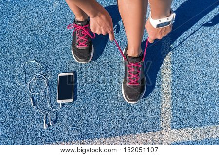 Runner woman tying running shoes laces getting ready for race on run track with smartphone and earphones for music listening on mobile phone. Athlete preparing for cardio training. Feet on ground.