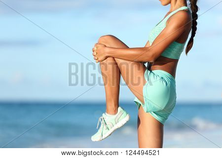 Fitness runner body closeup doing warm-up routine on beach before running, stretching leg muscles with standing single knee to chest stretch. Female athlete preparing legs for cardio workout.