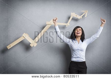 Concept: Success in business or career. Enthusiastic businesswoman with raised arms cheering in front of positive business graph, isolated on grey background.