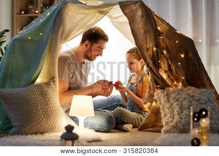 family, hygge and people concept - happy father and little daughter playing tea party in kids tent at night at home